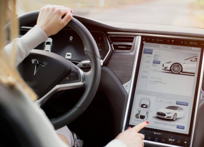 Interior de um veículo elétrico Tesla Model S; mulher dirigindo e tocando no painel touchscreen.