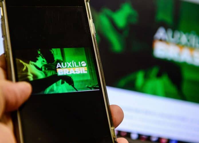 Pessoa segurando celular em frente ao computador, ambos com imagens do auxílio emergencial