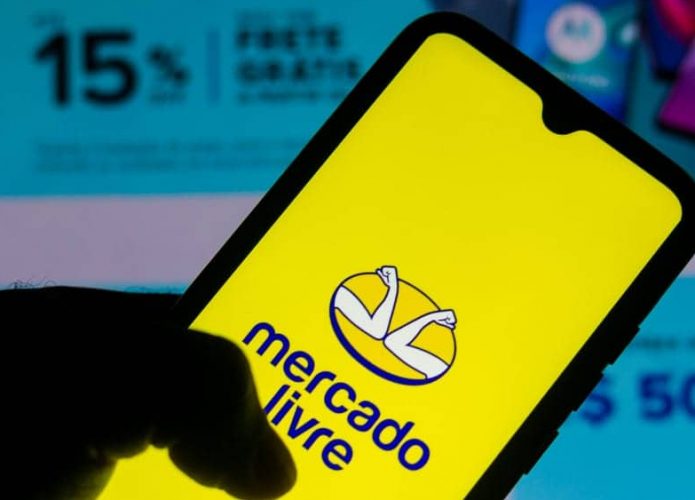 Pessoa segurando celular com logomarca do Mercado Livre na tela, colocado na frente de monitor exibindo promoções da varejista