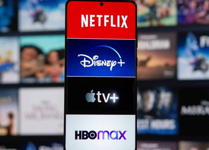 Celular com logomarcas de plataformas de streaming na tela e, ao fundo, estão imagens de produções disponíveis no streaming