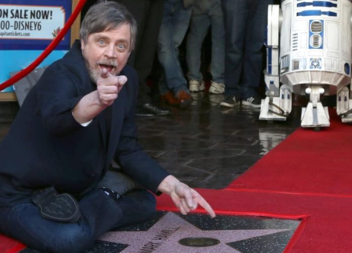 Mark Hamill sentado na calçada da fama com R2-D2 no fundo