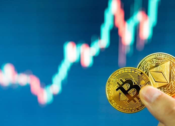 Pessoa segurando duas criptomoedas com linha de ações financeiras ao fundo