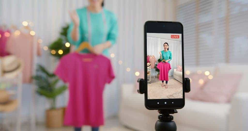 Mulher segurando blusa enquanto faz live; celular está num tripé