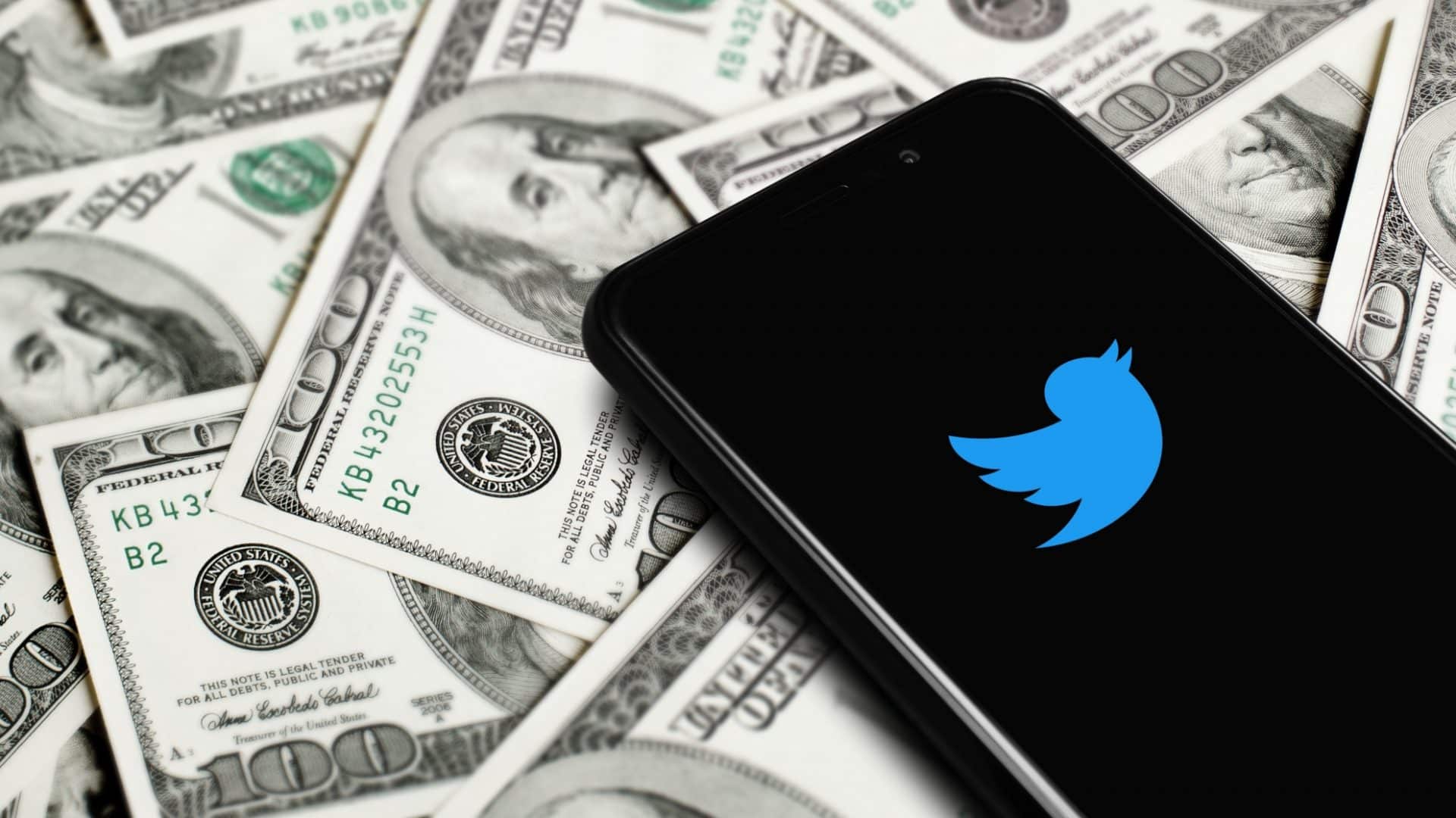 Logotipo azul do Twitter na tela de um smartphone com notas de dólares ao fundo. O Twitter é uma rede social comprada em 2022 por Elon Musk.