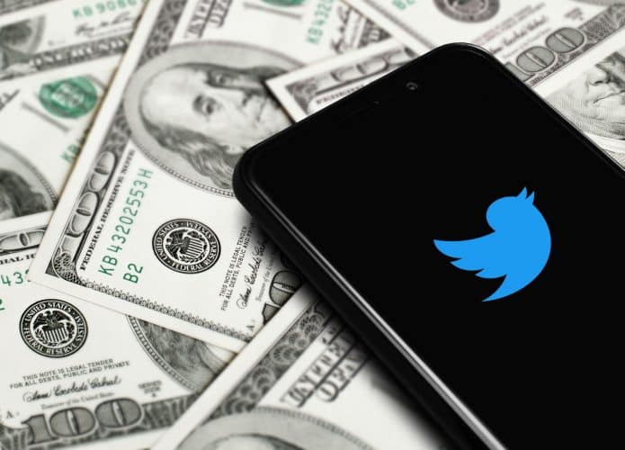 Logotipo azul do Twitter na tela de um smartphone com notas de dólares ao fundo. O Twitter é uma rede social comprada em 2022 por Elon Musk.