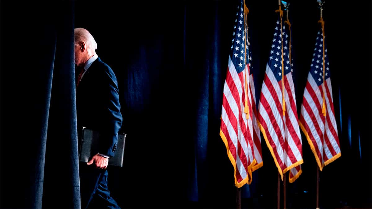 Joe Biden saindo de palco, com rosto ocultado na coxia e três bandeiras dos EUA ao fundo