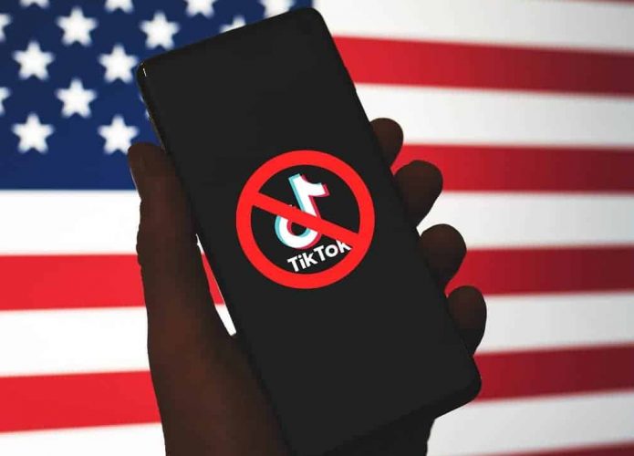 Pessoa segurando celular com logotipo do TikTokna tela e sinal de bloqueio em cima; ao fundo, a bandeira dos Estados Unidos