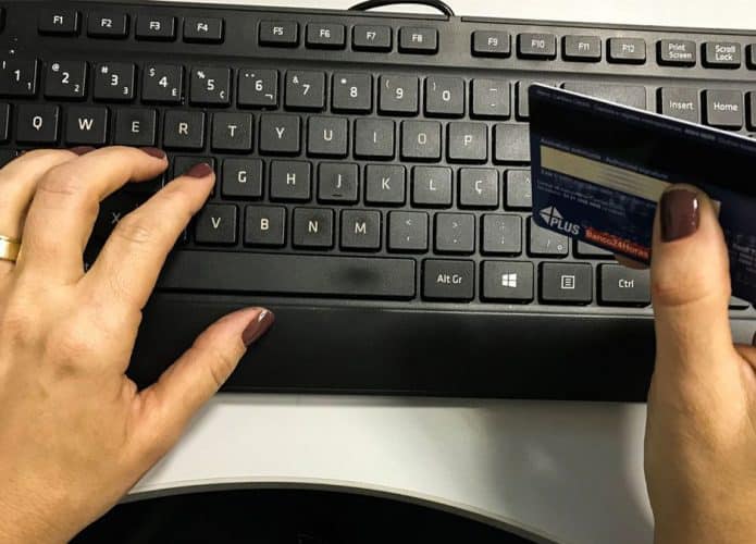 Mulher com uma mão sobre teclado e outra segurando cartão de débito e crédito