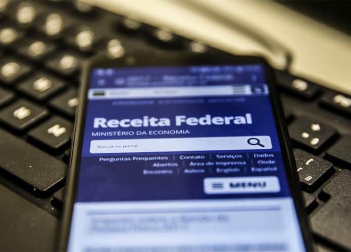 Celular com homepage da Receita Federal aberta em navegador; aparelho está sobre teclado de computador