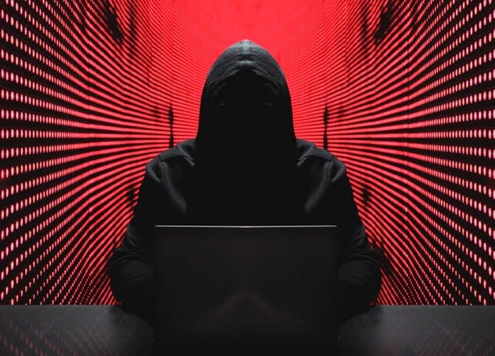 Hacker encapuzado sentado usando notebook numa mesa e, ao fundo, paredes com luzes vermelhas