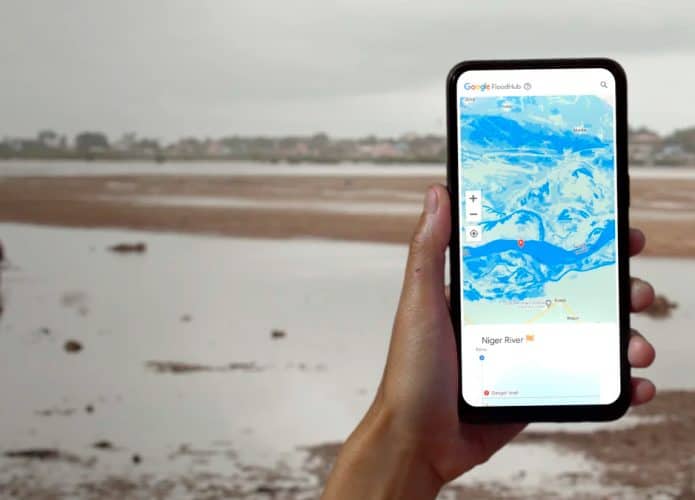 Pessoa segurando celular com Flood Hub aberto e, ao fundo, paisagem com enchente ribeirinha