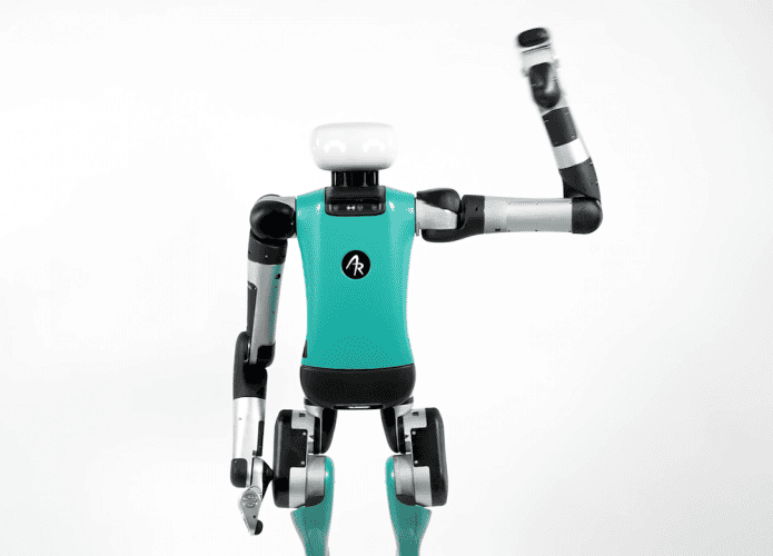 A imagem mostra o robô Digit, da Agility Robotics. Ele é um dispositivo robótico nas cores verde água, cinza e preto, e tem a cabeça branca. Na foto, ele aparece com a mão esquerda levantada, como se fizesse uma saudação.