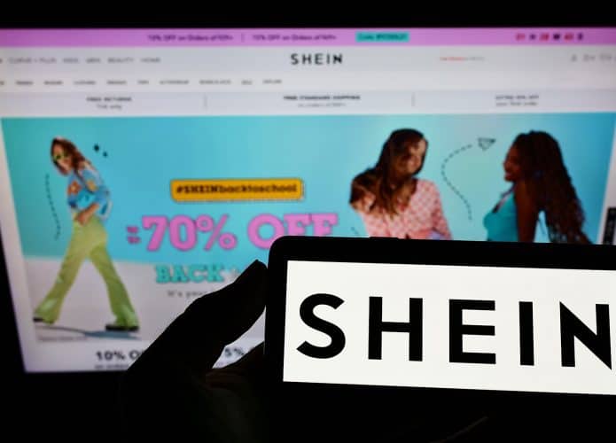 Pessoa segurando celular com logotipo da Shein na tela enquanto a homepage do site está aberta num computador ao fundo