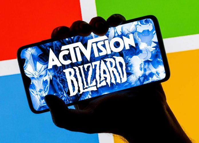 Pessoa segurando celular com imagem da logomarca da Activision na tela e, ao fundo, logomarca do Windows, da Microsoft