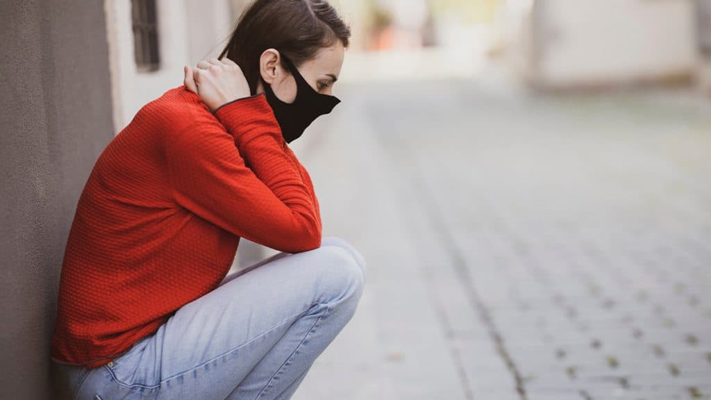 Mulher agachada, usando máscara e com mão na nuca, sentindo sintomas de Covid