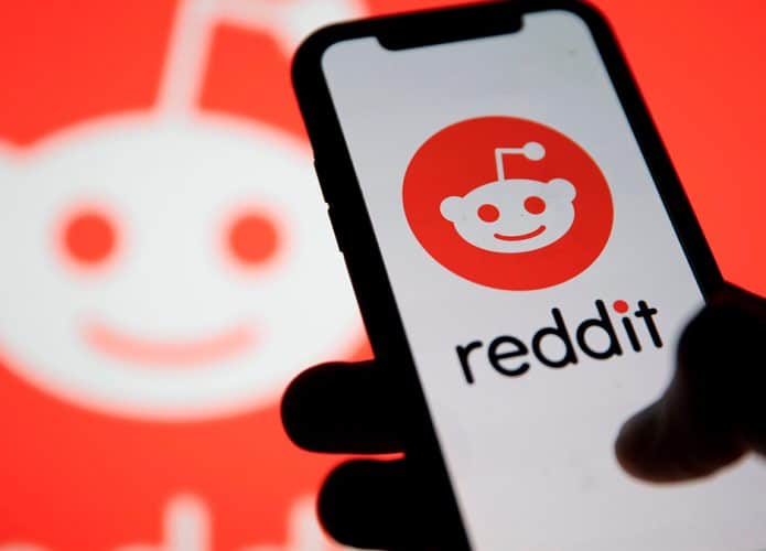 Pessoa segurando iPhone com logomarca do Reddit na tela e, ao fundo, logomarca do Reddit em outra tela