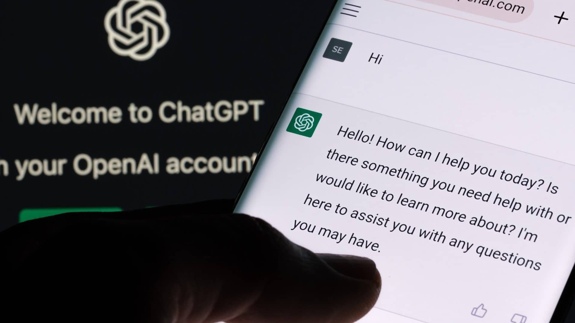 Pessoa com dedo sobre conversa no ChatGPT no celular com chatbot aberto num computador ao fundo