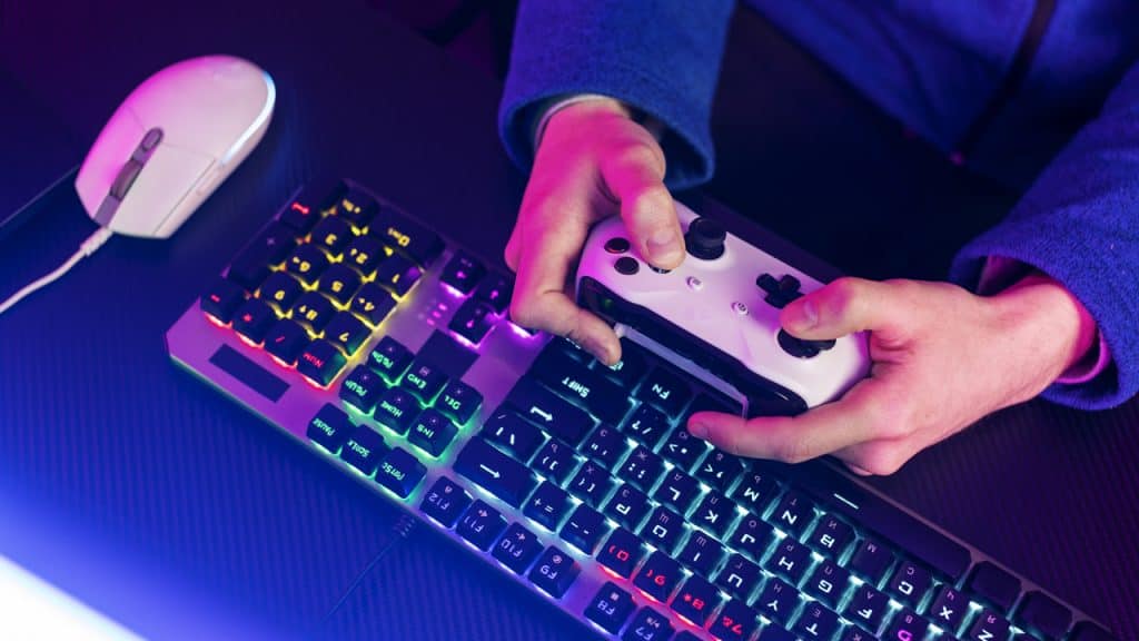 pessoa com controle de videogame na mão numa mesa com teclado e mouse