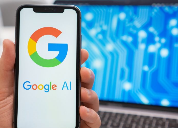 Pessoa segurando celular com logomarca da IA do Google na tela e, ao fundo, um notebook com imagem ilustrativa de tecnologia na tela
