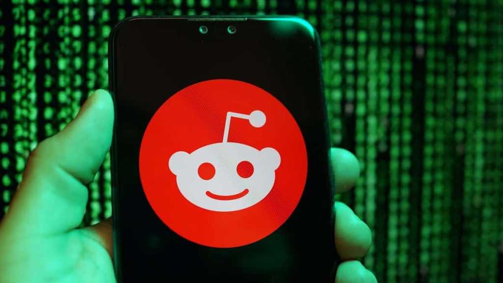 Pessoa segurando celular com a logomarca do Reddit na tela e, ao fundo, aparecem linhas de programação