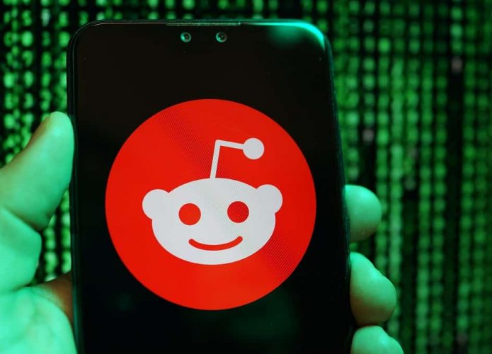 Pessoa segurando celular com a logomarca do Reddit na tela e, ao fundo, aparecem linhas de programação