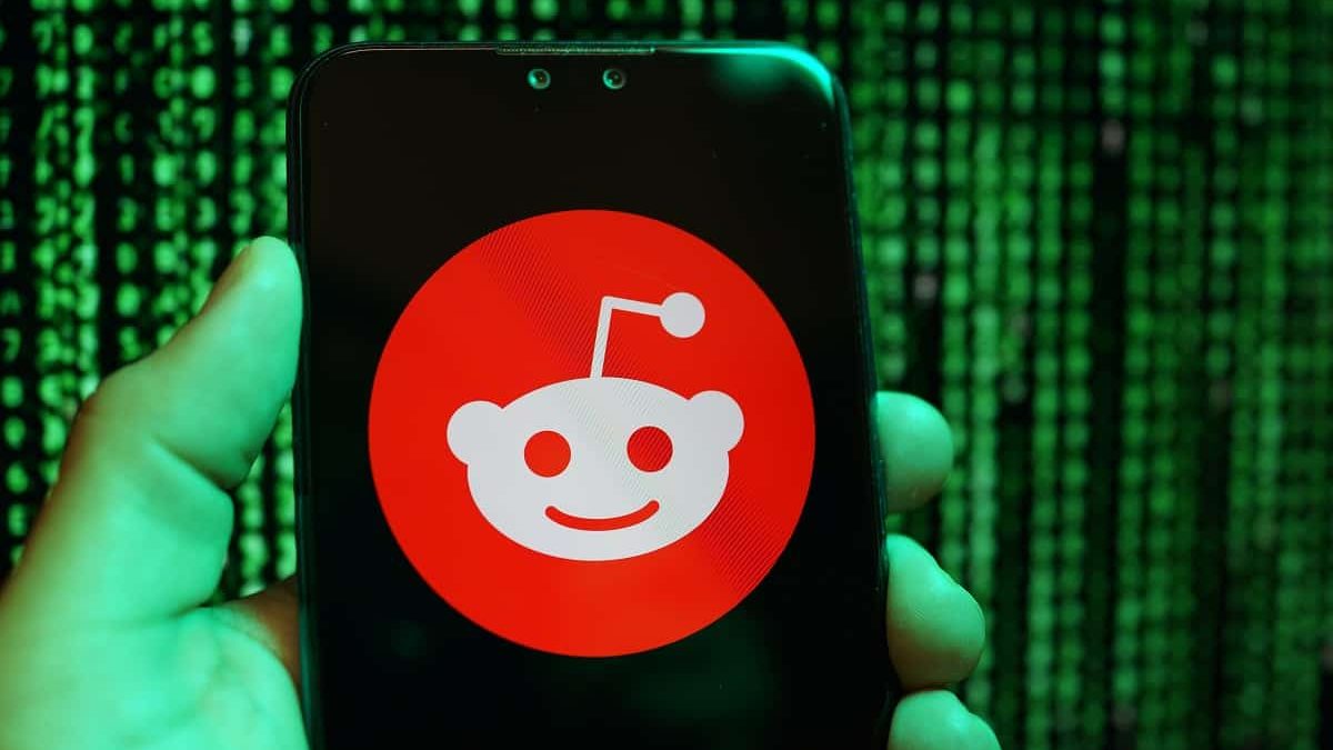 Pessoa segurando celular com a logomarca do Reddit na tela e, ao fundo, aparecem linhas de programação