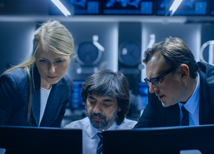 Uma mulher e dois homens encarando duas telas de computadores, á sua frente.