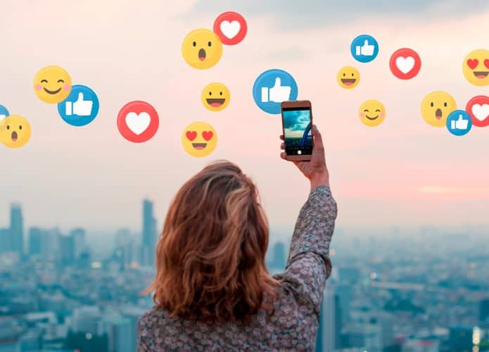 Mulher tirando uma selfie no pôr do sol com ícones de reações do Facebook no ar