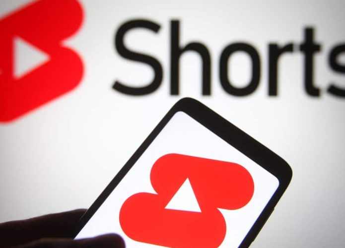 Pessoa segurando celular com logo do YouTube Shorts na tela, também exibido numa tela ao fundo