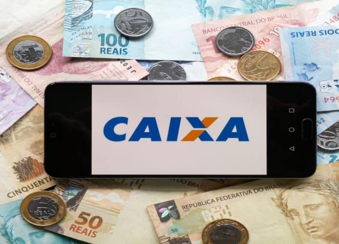 Celular com logo da caixa na tela e dinheiro em real de notas e moedas