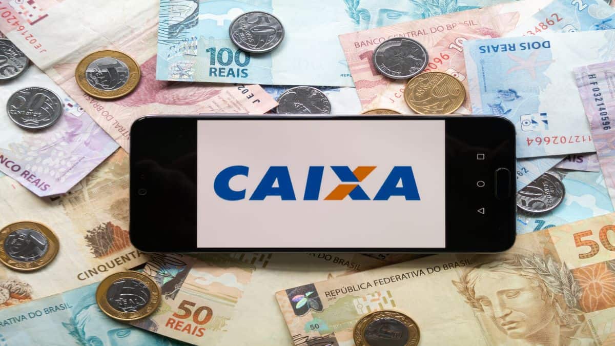 Celular com logo da caixa na tela e dinheiro em real de notas e moedas