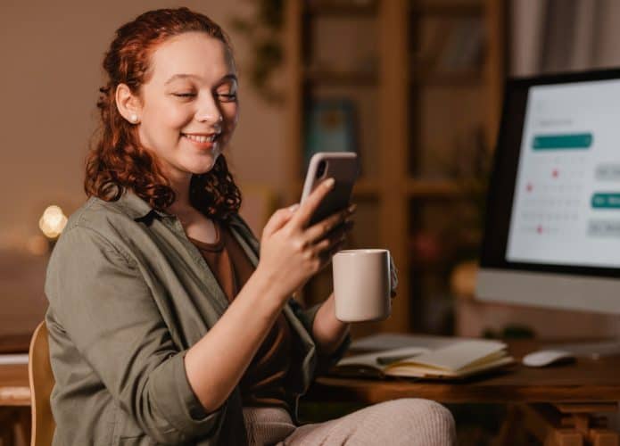 mulher-em-casa-usando-smartphone-na-frente-do-computador-enquanto-toma-cafe-695x500