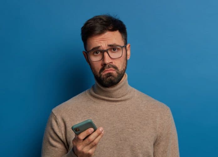 barbudo-descontente-com-um-sorriso-afetado-usa-um-celular-moderno-tem-uma-expressao-triste-usa-oculos-transparentes-e-macacao-695x500