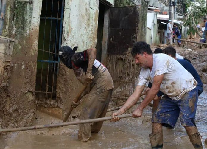 Homens tirando lama de fachada de casa e rua após desastre natural em morro