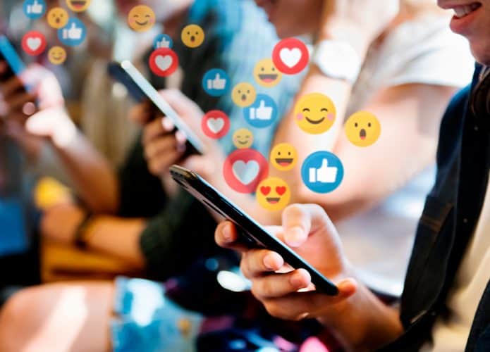 Jovens usando celular com emojis de reações de redes sociais saindo dos aparelhos