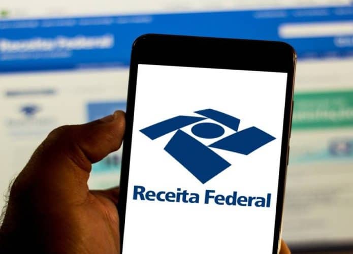 Tela de navegador no site da Receita Federal de fundo e celular em primeiro plano com logo