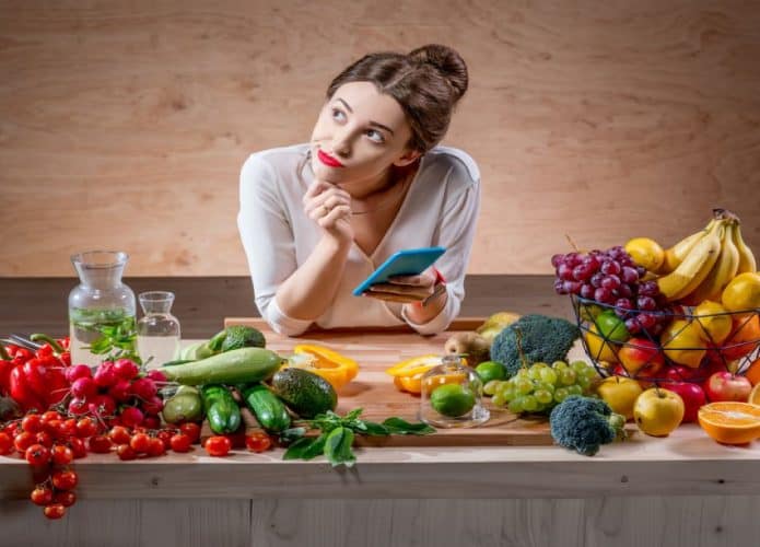 imagem de uma mulher com cara de dúvidas e segurando celular em uma das mãos e a mesa repleta de frutas, verduras e legumes