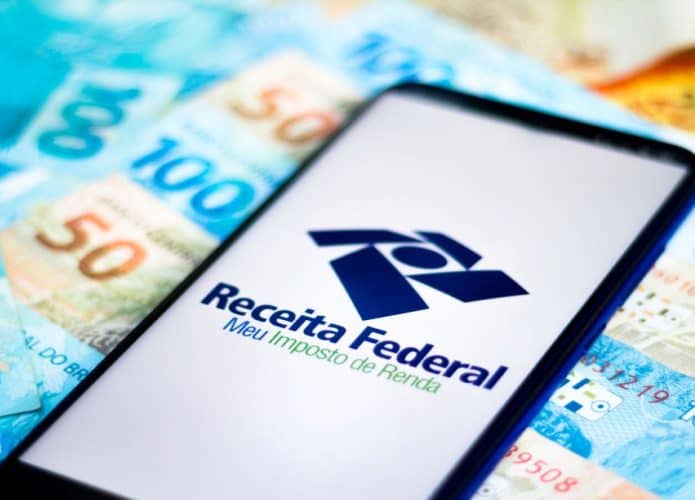 notas de cem reais e cinquenta e celular com logo da Receita Federal na tela