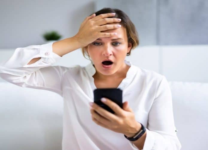 mulher com expressão de susto com boca aberta e mão na testa olhando para tela de celular