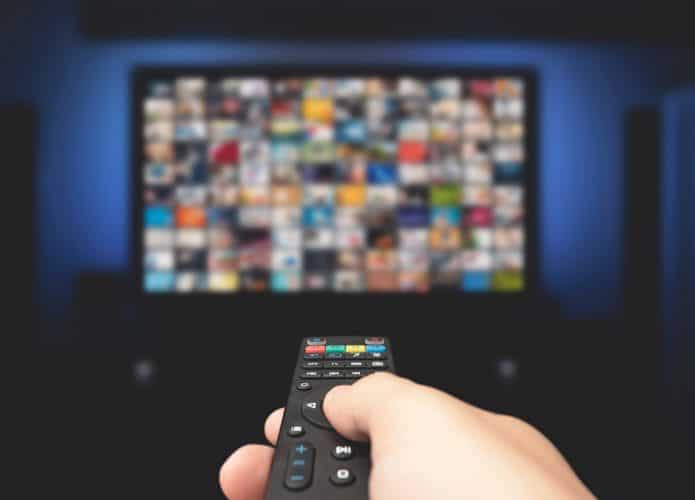 Pessoa apontando controle para TV lotada de imagens de programas; imagem ilustra conceito de TV 3.0