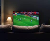Sofá de costas; à frente, uma TV com um jogo de futebol sendo transmitido