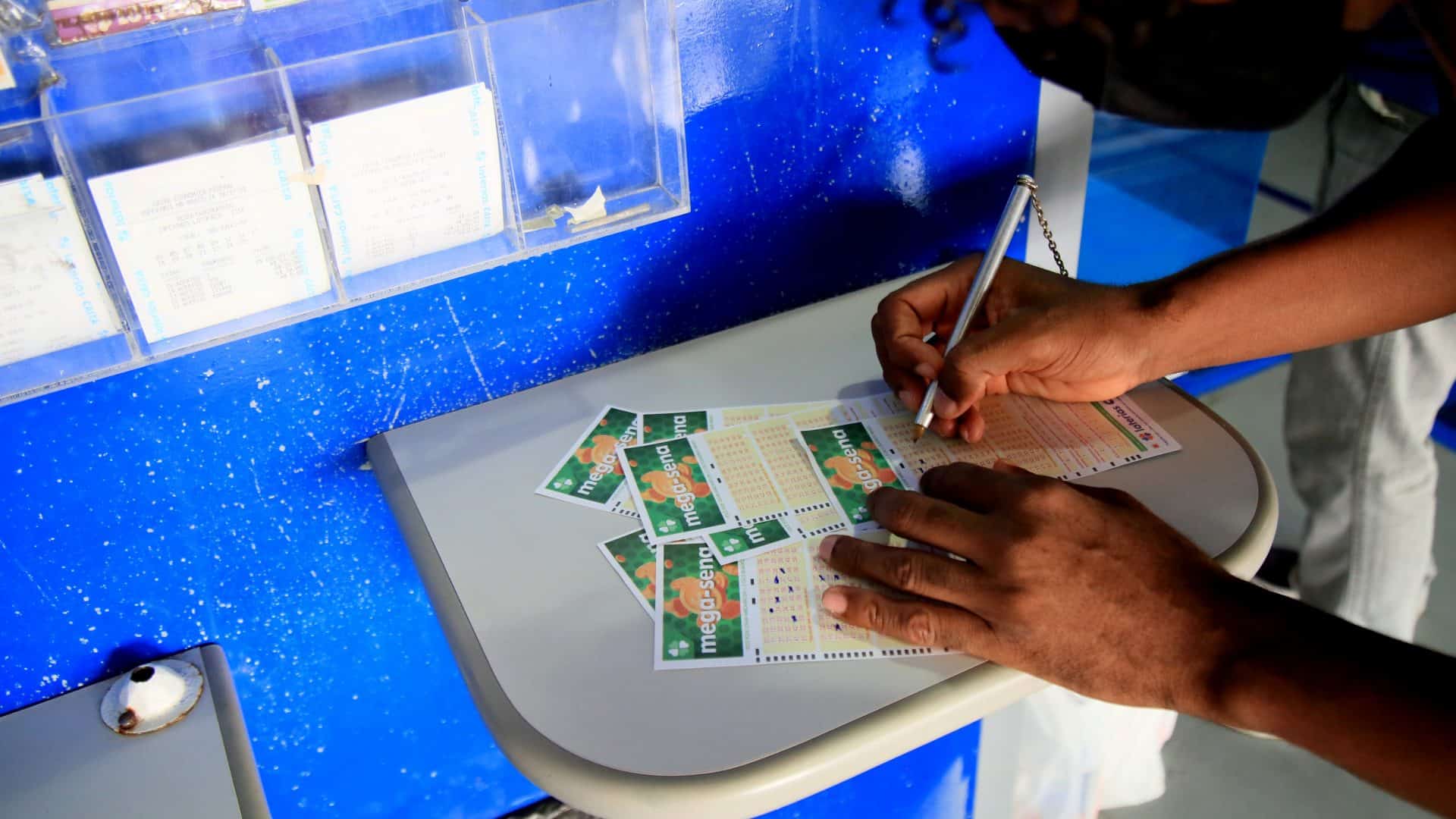 Pessoa preenchendo volantes da mega-sena em cima de um balcão de uma lotérica