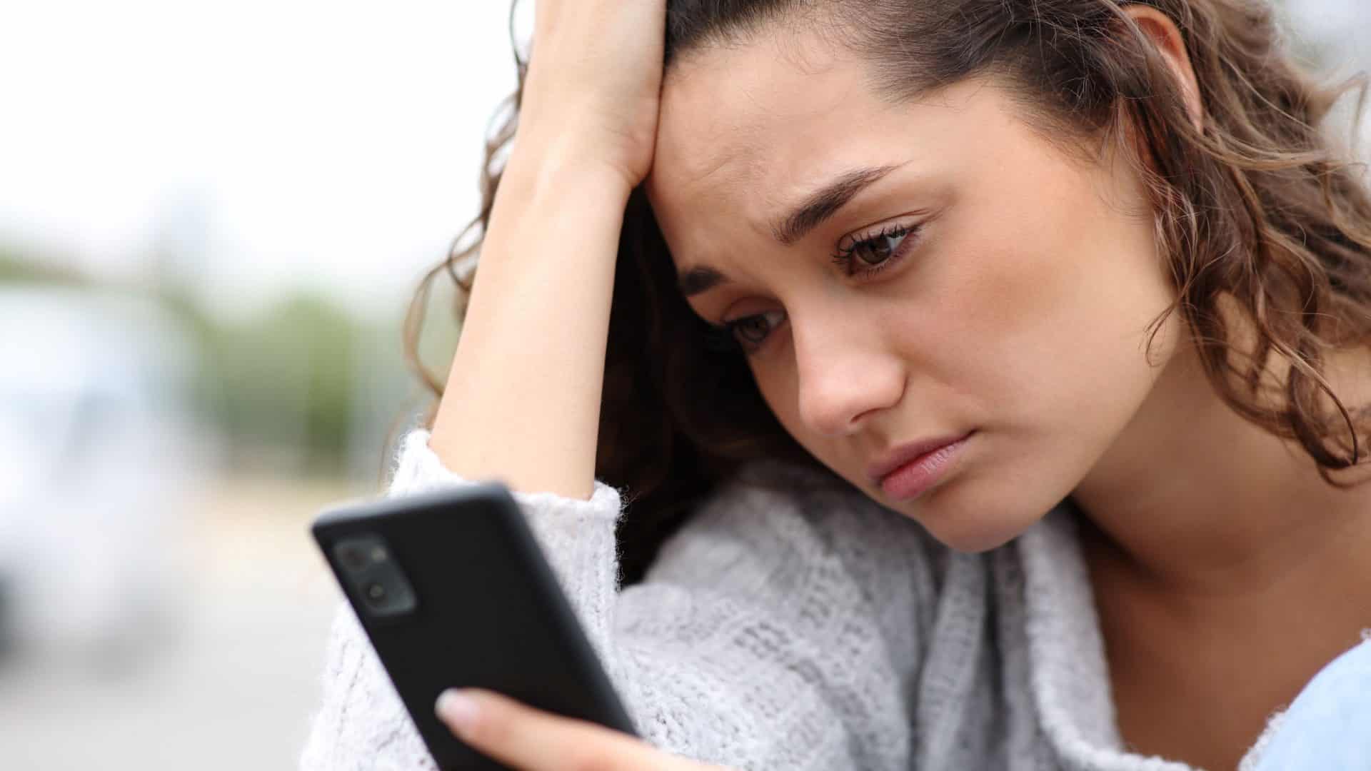 mulher com cara de triste com uma mão segurando o celular e a outra na cabeça