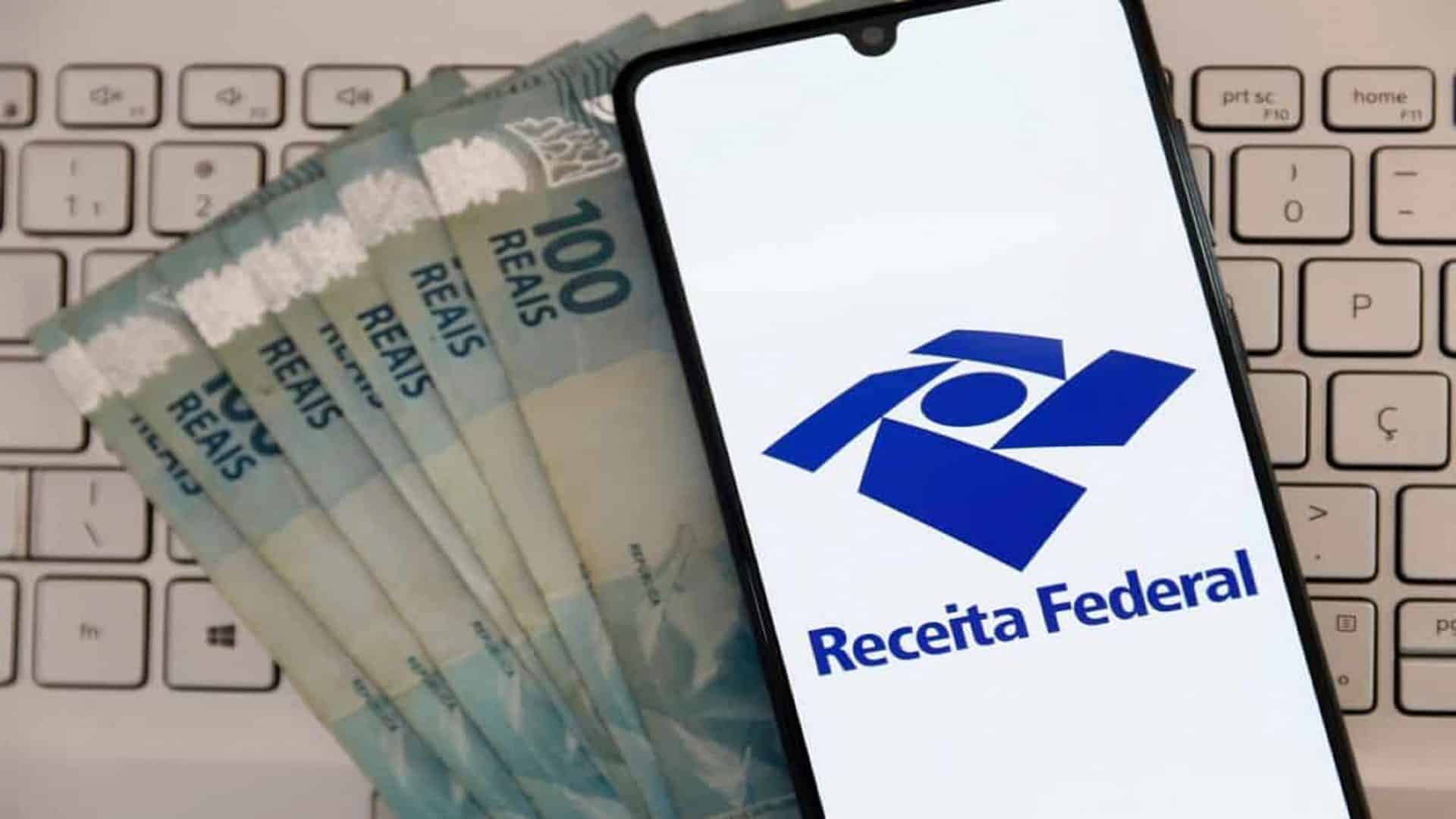 Celular com logotipo da Receita Federal em cima de notas de 100 reais e teclado de notebook