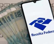Celular com logotipo da Receita Federal em cima de notas de 100 reais e teclado de notebook