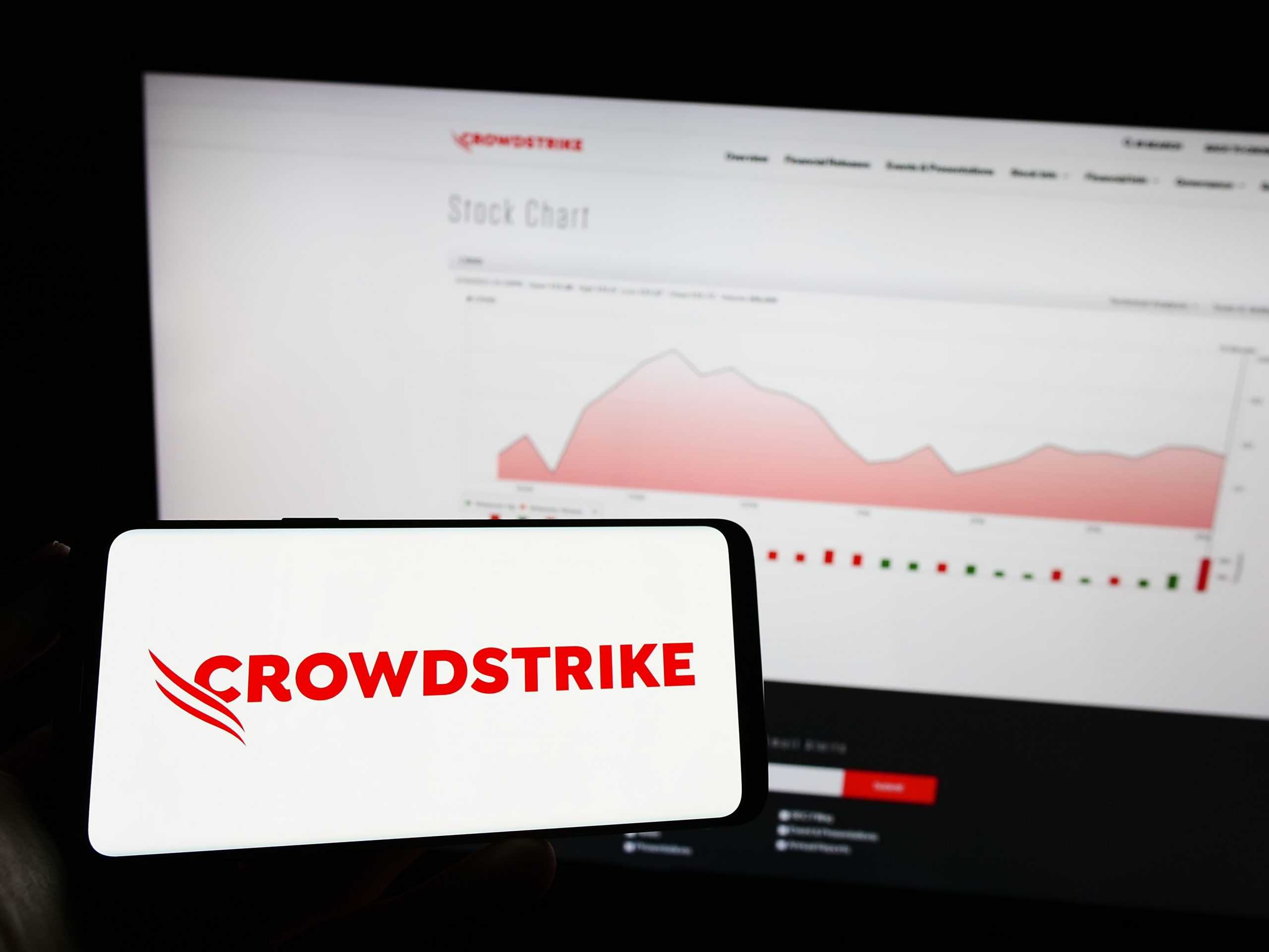 Logo da CrowdStrike em smartphone, com fundo de um monitor mostrando um gráfico no site da empresa