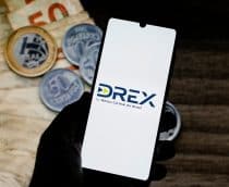 Imagem mostra, ao fundo, em uma mesa, notas de real e moedas; à frente, um smartphone com o logo do Drex na tela