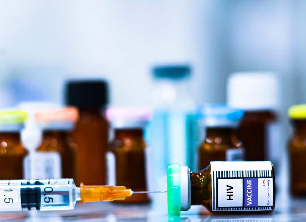 Vários medicamentos ao fundo, com uma seringa injetada em um medicamento à frente, no qual se lê "HIV"