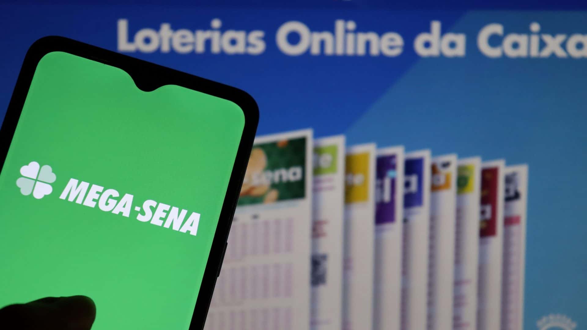 Logo da Mega-Sena em celular à esquerda com fundo desfocado, no qual há uma propaganda da Loterias Caixa