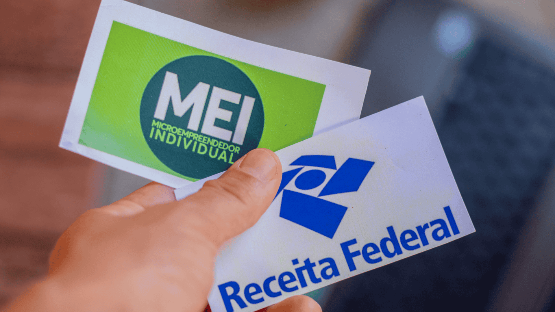 imagem demonstra uma mão segurando duas fichas: a primeira tem a estampa do MEI, e a segunda a estampa da receita federal